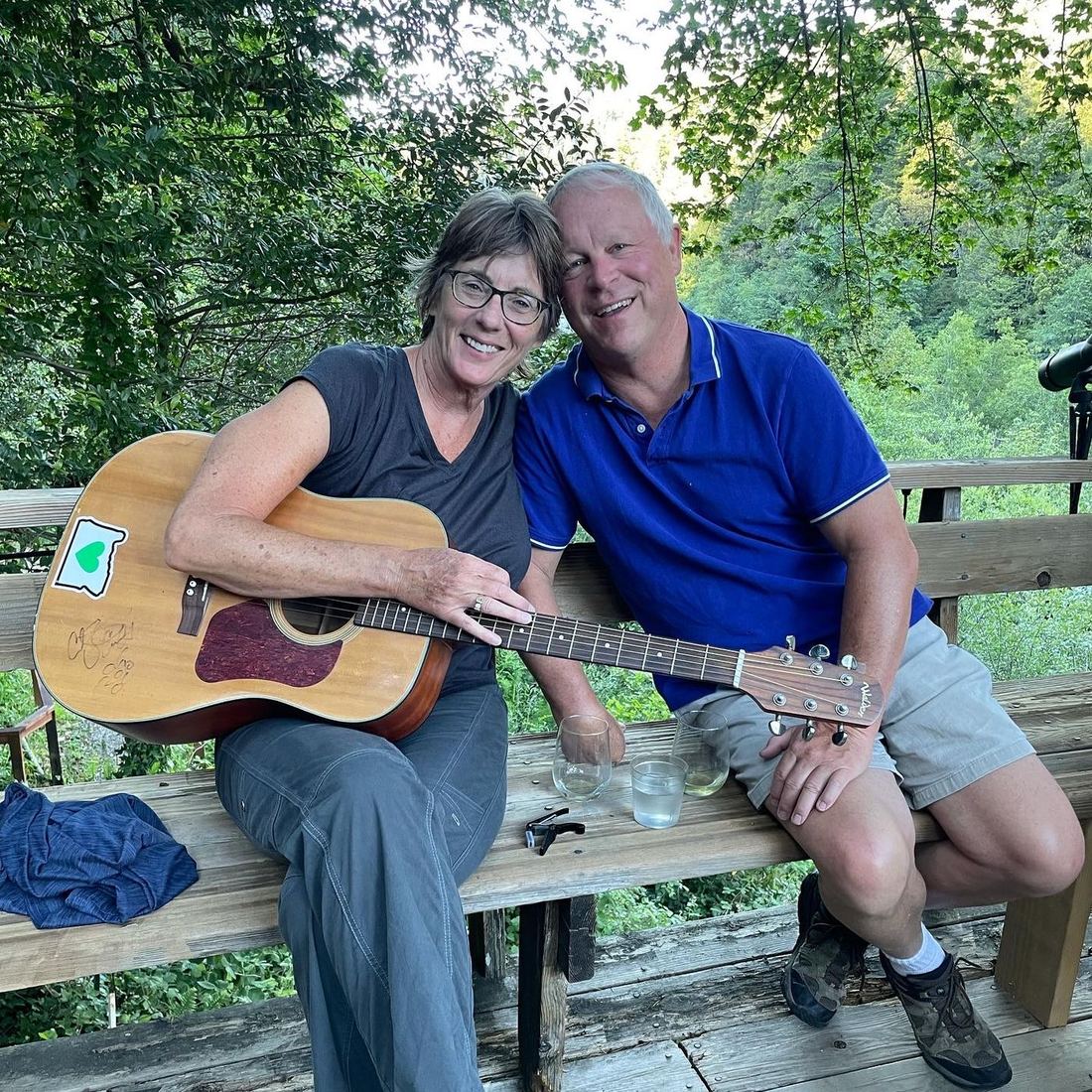 Terry and Liz McAteer sitting together on a wooden deck in the trees, Liz holding a guitar